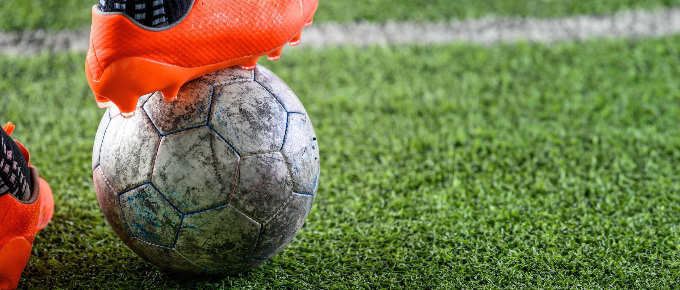 Worn soccer ball on green grass field with bright orange cleats stepping on it, showing action and sports energy in close up shot photo