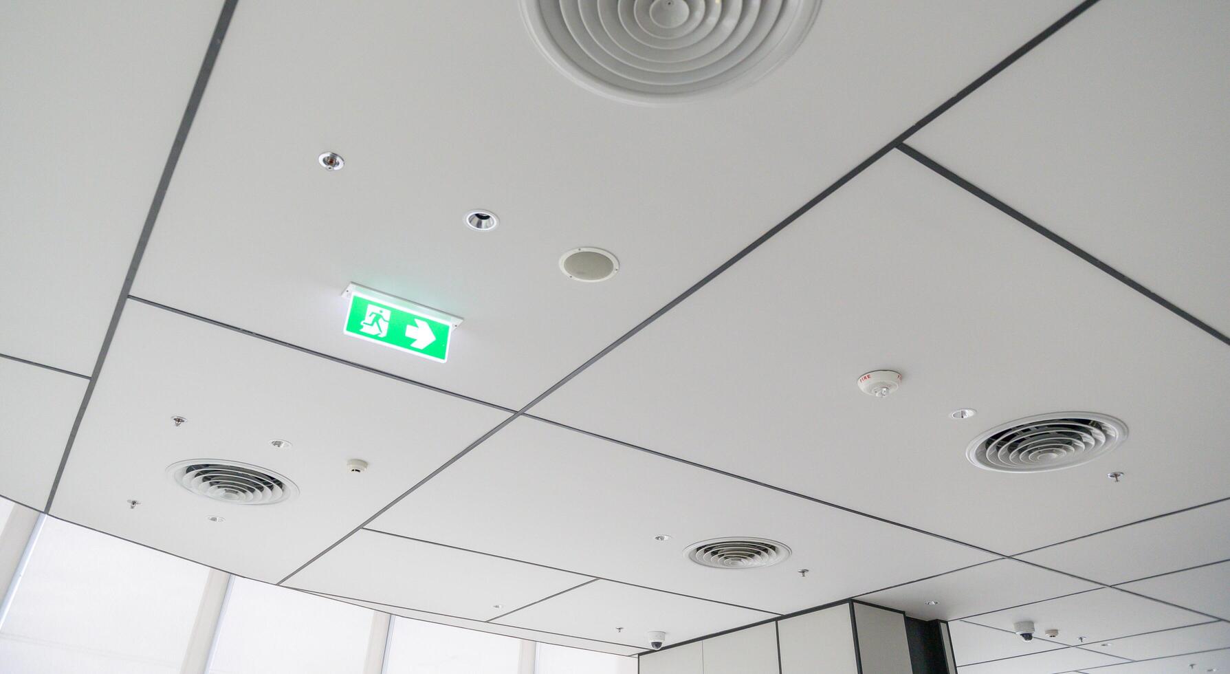 Clean modern office ceiling featuring air vents, recessed lighting, and grid pattern with illuminated emergency exit sign, creating bright and organized atmosphere photo