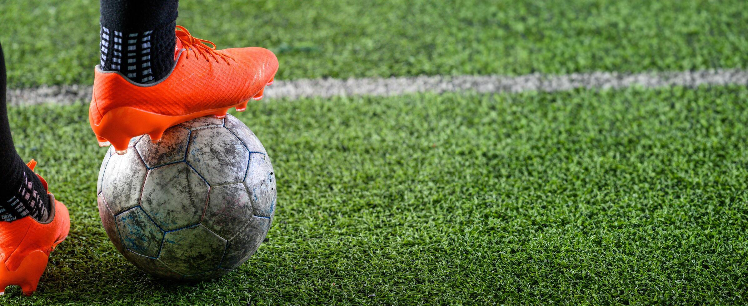 Worn soccer ball on green grass field with person wearing bright orange cleats standing on it, showing sports action and outdoor activity in close up shot photo