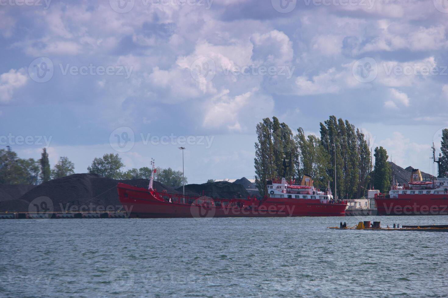 Red cargo ships docked at a busy industrial port with coal piles and trees in the background photo
