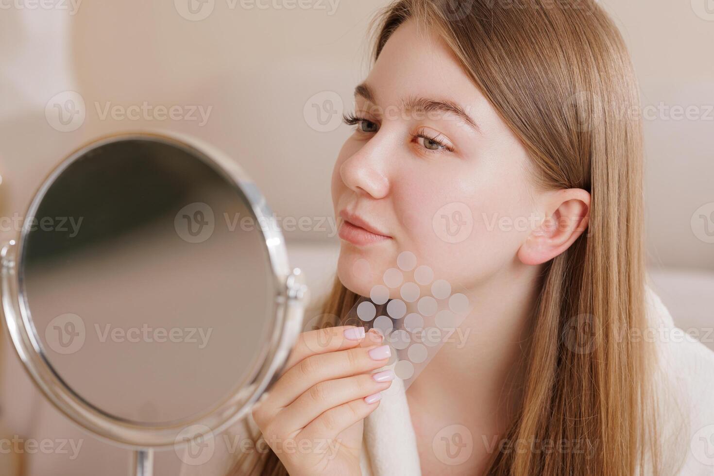 Girl examines face in mirror while holding pimple patch sheet for skincare treatment. photo