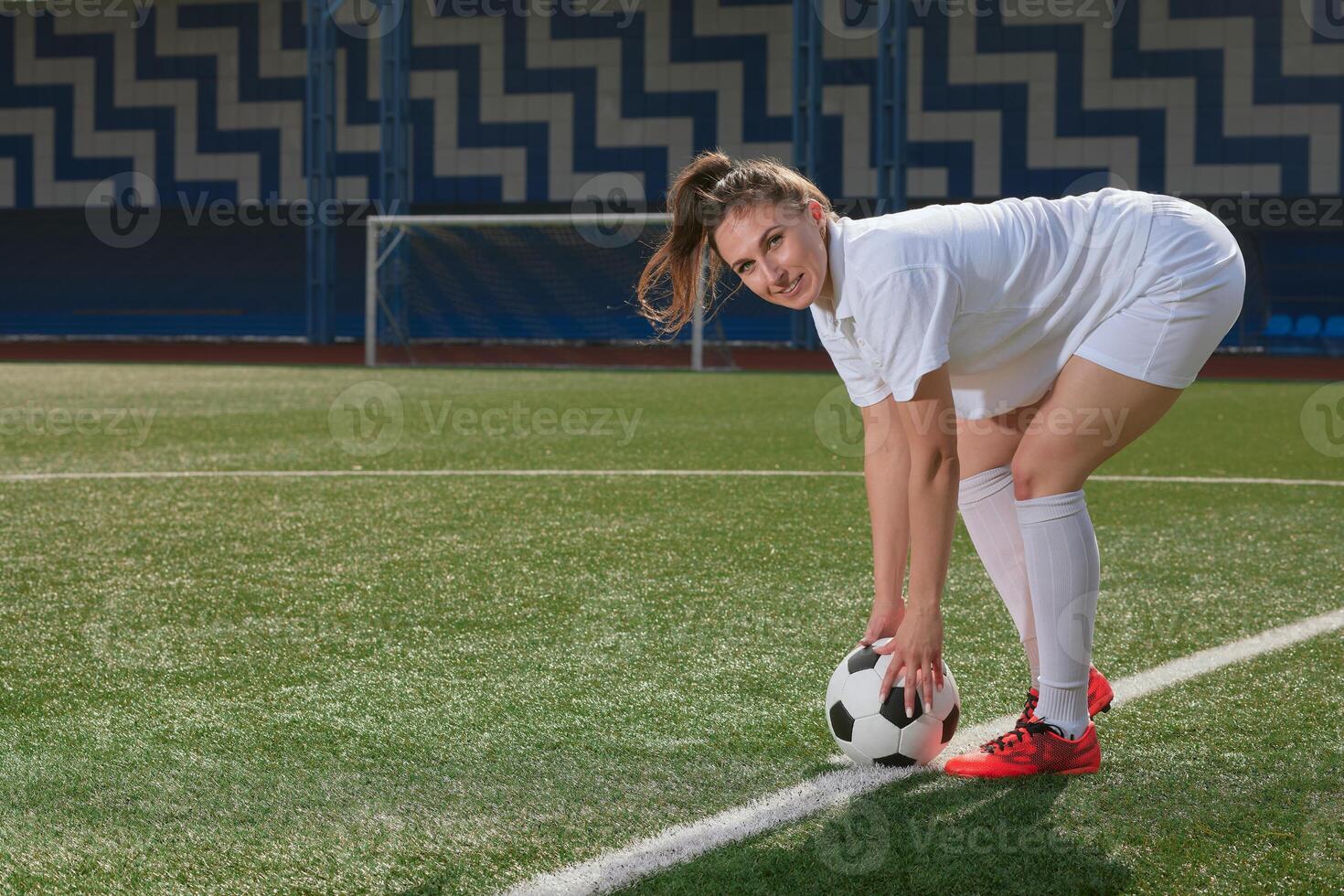 Smiling woman football player in white sportswear holding soccer ball on green field. photo