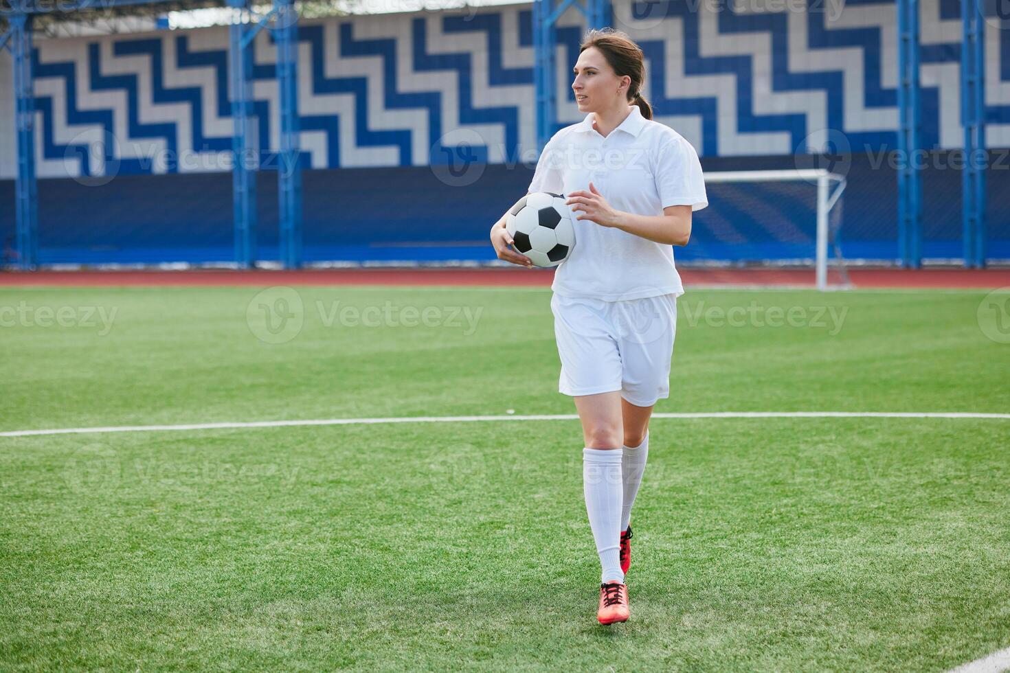 Woman in soccer uniform holding ball, walking confidently on green field. photo