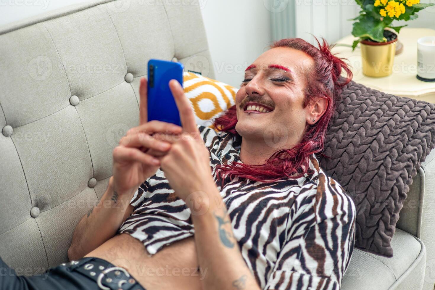 Non-binary person with pink hair using smartphone and smiling on sofa photo