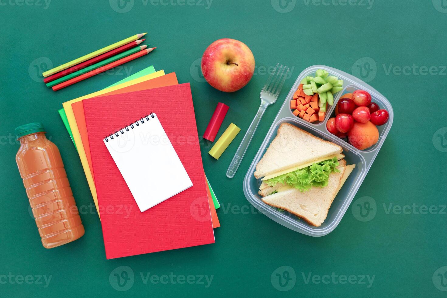 Colorful arrangement of lunch items includes sandwich with lettuce, assorted fruits, juice bottle, and stationery on green surface ready for midday meal. photo
