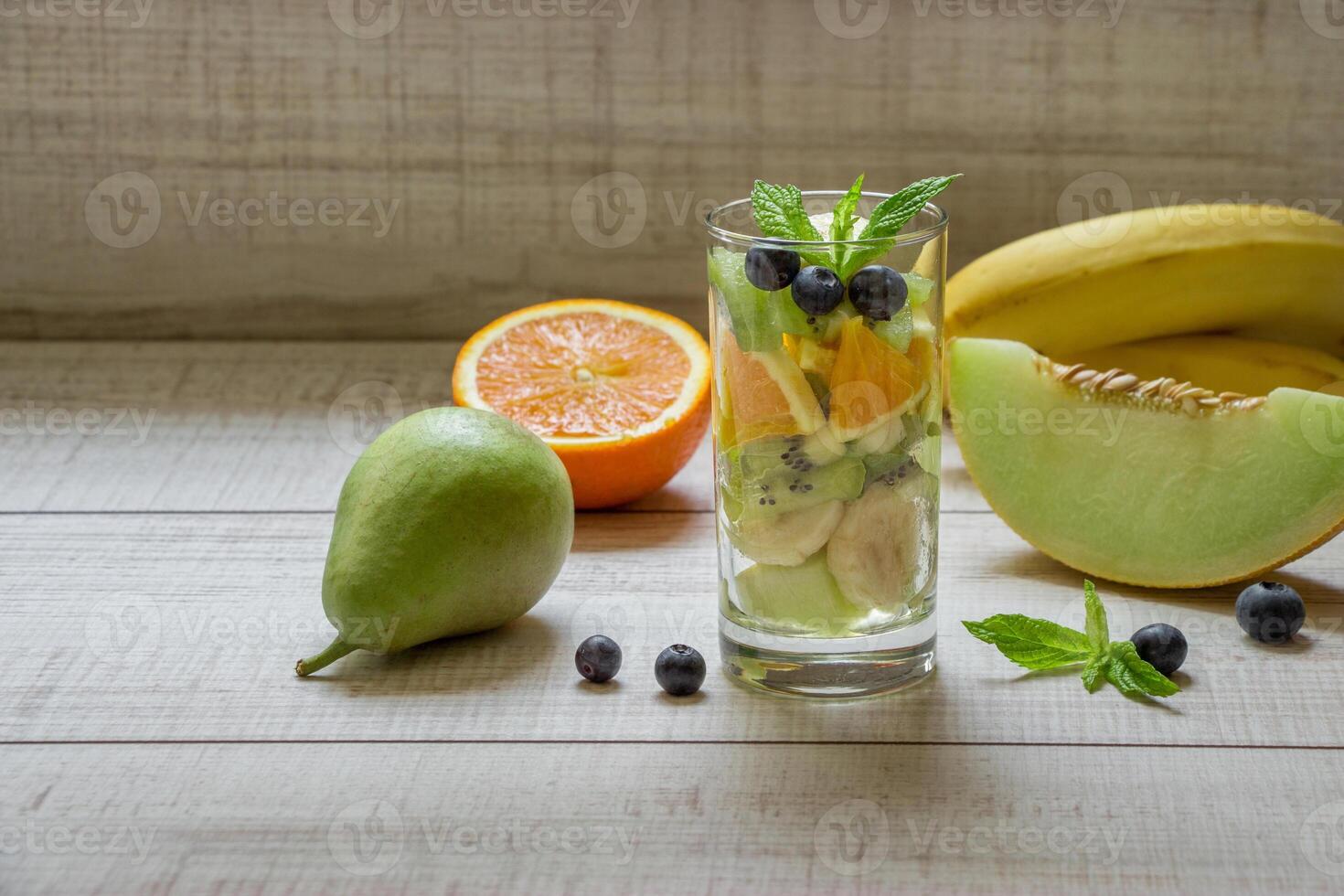 Right glass with sliced fruits melon, banana, orange, kiwi, pear next to ingredients, left empty space for text also in frame blueberries, mint on light background. Glass of fruit salad. Horizontal. photo