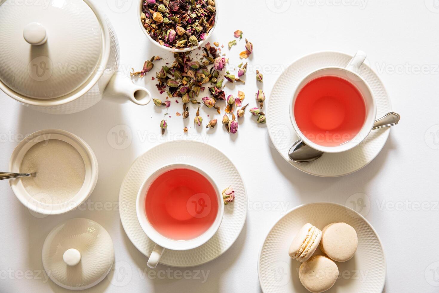 Two cups filled with rose infused tea sit beside delicate macarons and a bowl of dried flower petals on a clean white table. A teapot and spoon complete the inviting setup. photo