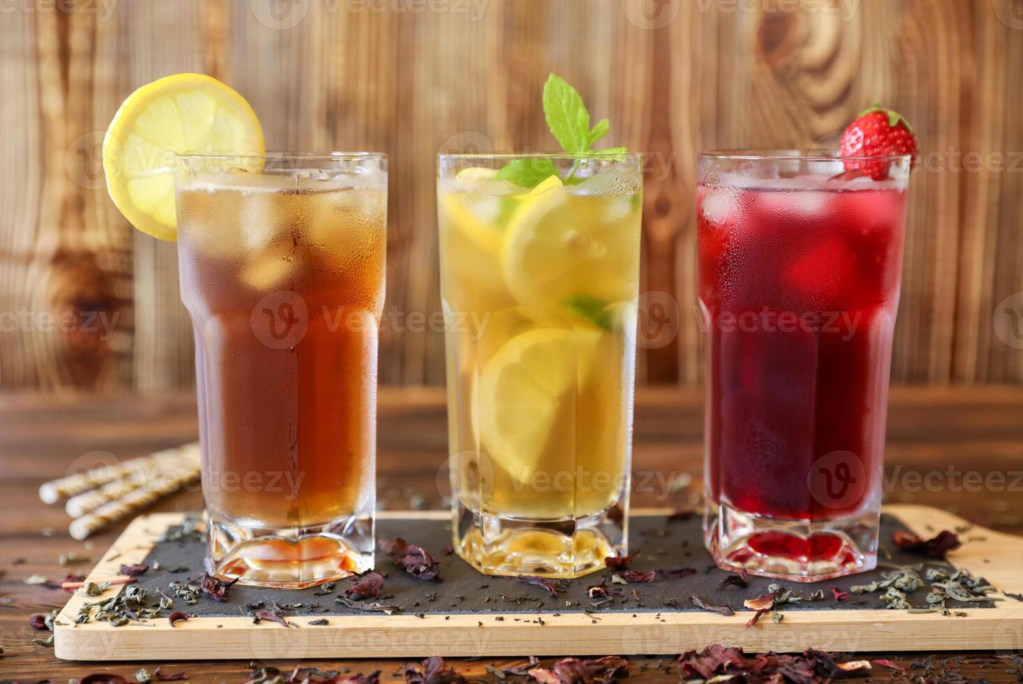Three tall glasses filled with iced tea, lemonade with lemon slices, and a berry drink with cherries are presented on rustic wooden table. Fresh mint garnish adds vibrant touch. photo