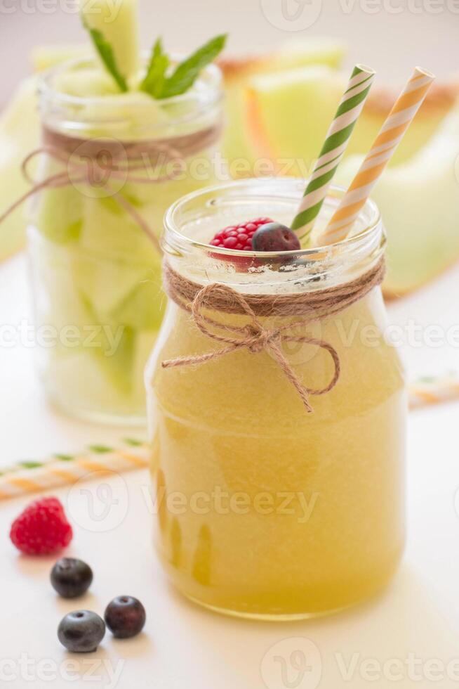 Jar with melon smoothie near berries, straws diagonally behind a jar with melon pieces, melon cut slices on a white background. Melon smoothie and salad in 2 jars. Vertical. Close-up. photo