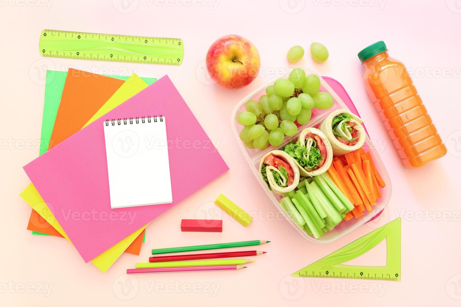 Healthy lunch features colorful wraps filled with vegetables, grapes, apple, and drink alongside bright stationery. Perfect setting for school or study time preparation. photo