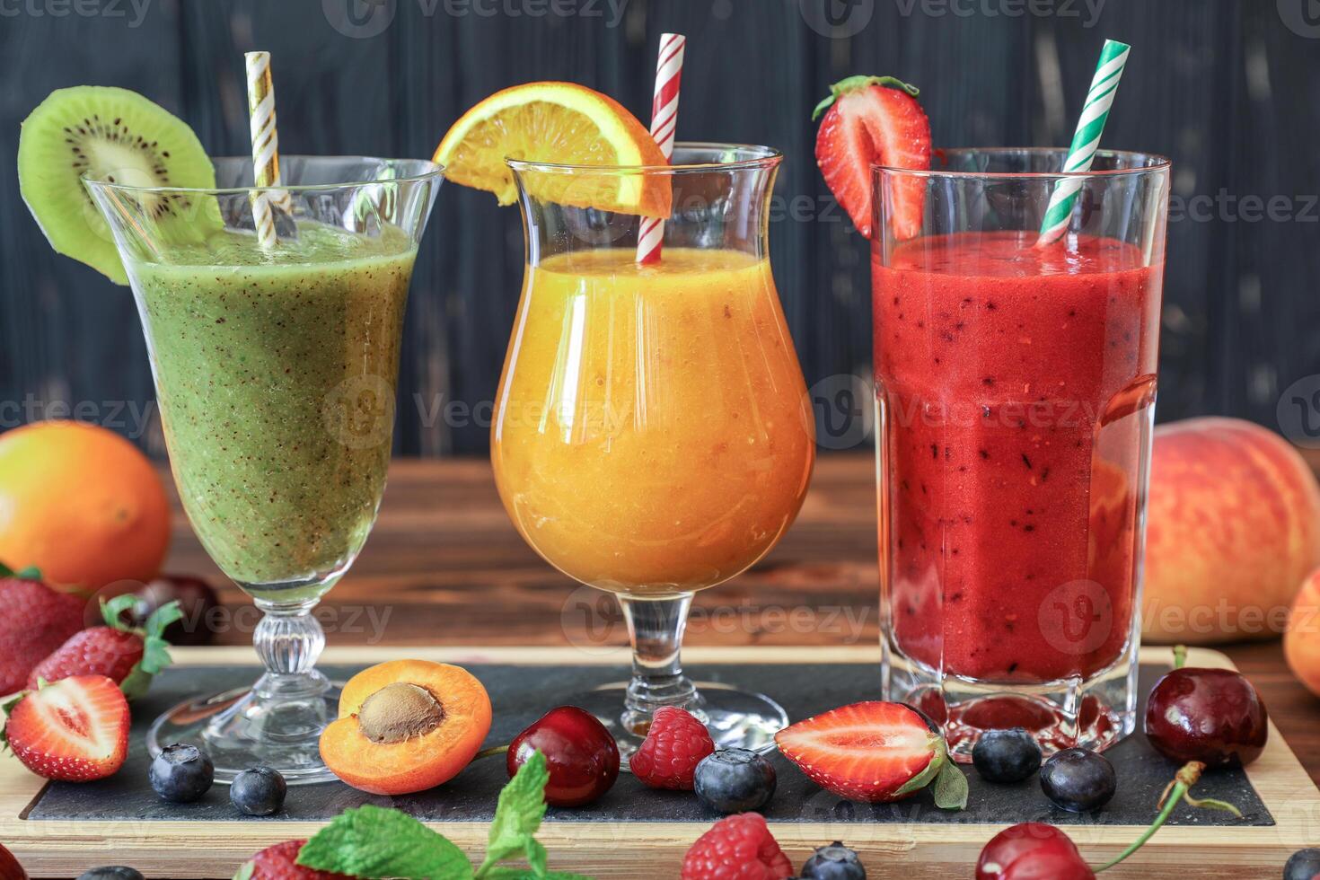 Three tall glasses filled with green, orange, and red smoothies are arranged on a wooden table, surrounded by fresh fruits like strawberries, kiwis, and cherries, perfect for hot days. photo