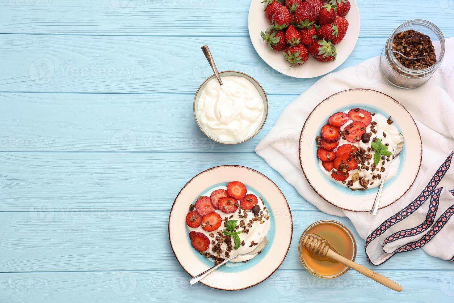 Two bowls filled with yogurt topped with sliced strawberries and granola sit on a light blue wooden table alongside honey, fresh strawberries, and utensils. photo