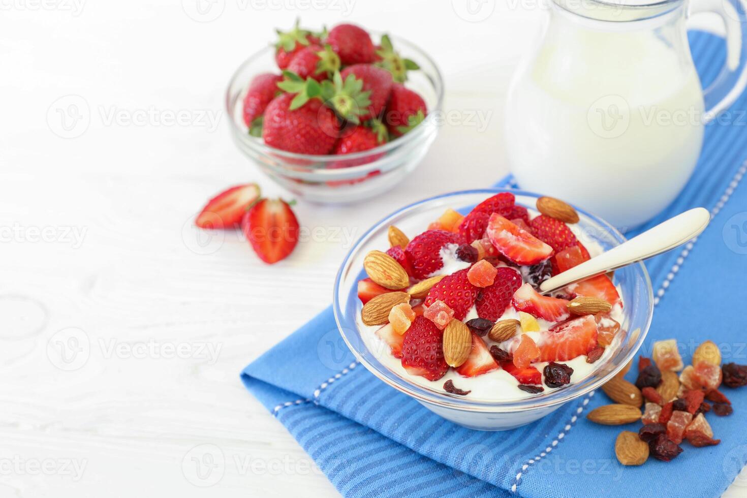 Bowl filled with creamy yogurt topped with ripe strawberries, almonds, and dried fruits sits on a blue cloth. Fresh strawberries and a jug of milk accompany it, enriching a healthy breakfast routine. photo