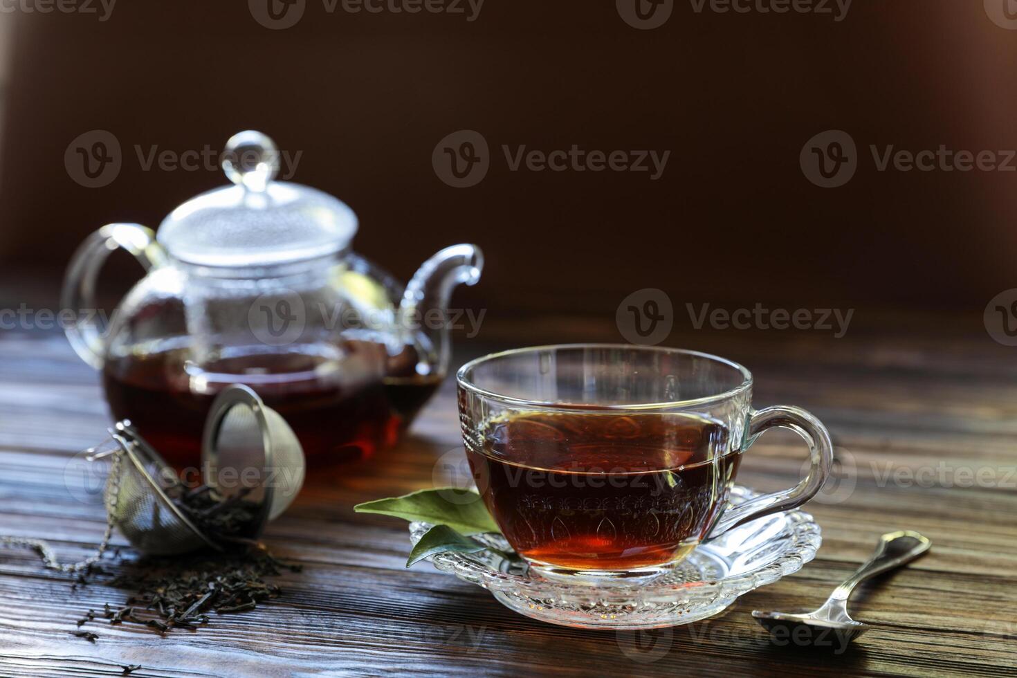 Brewed black tea sits in a crystal glass cup on a decorative saucer. Nearby, a transparent teapot holds more tea, and loose tea leaves lie scattered on the wooden table. photo