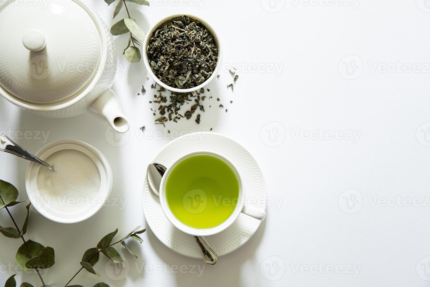 Bright green tea prepared in a simple cup alongside loose tea leaves, sugar bowl, and eucalyptus branches creating a calming atmosphere in a minimalist setting. photo