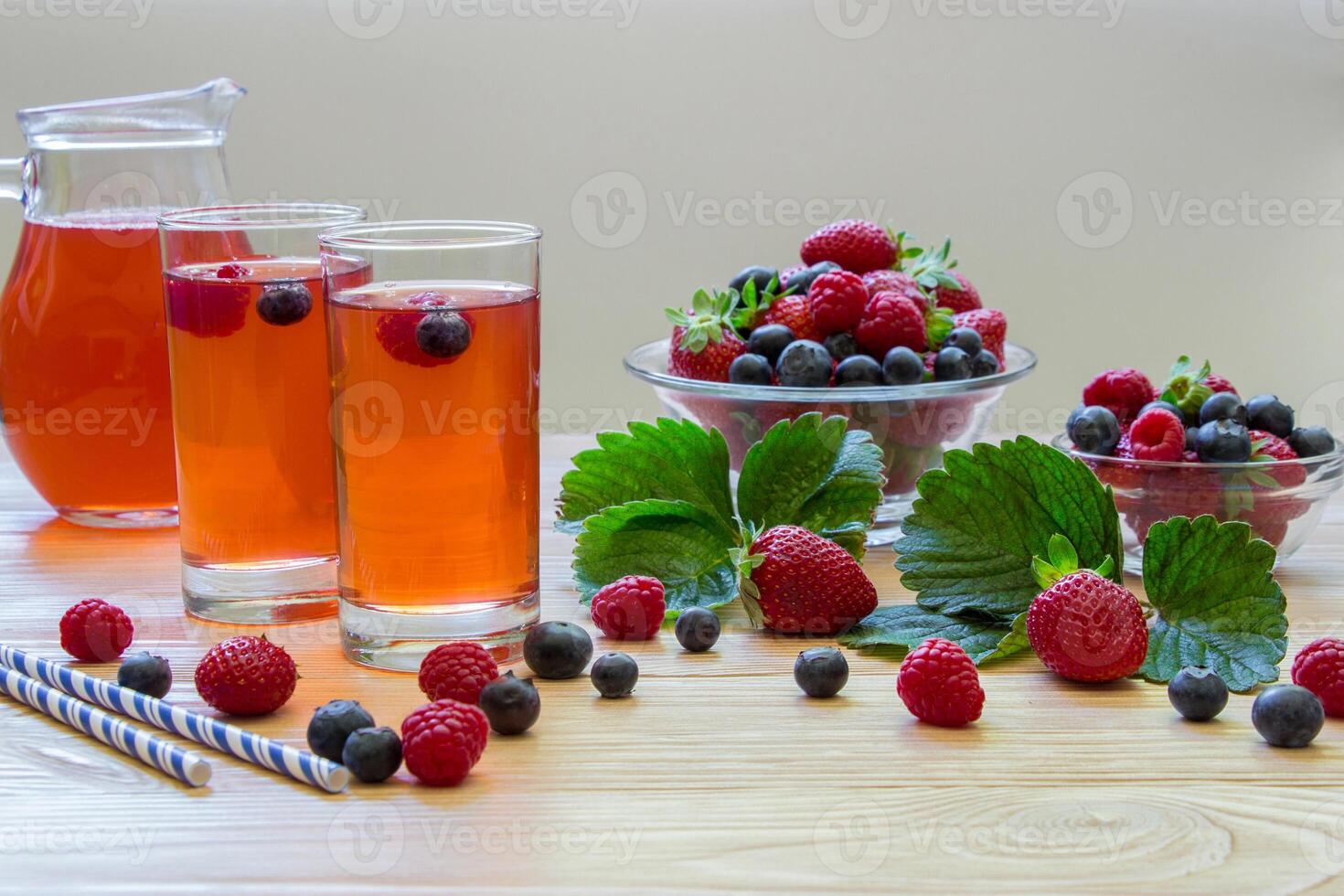 Two glasses filled with a berry beverage sit on a wooden table beside bowls of fresh raspberries and blueberries. Strawberries add a pop of color to the lively arrangement. photo