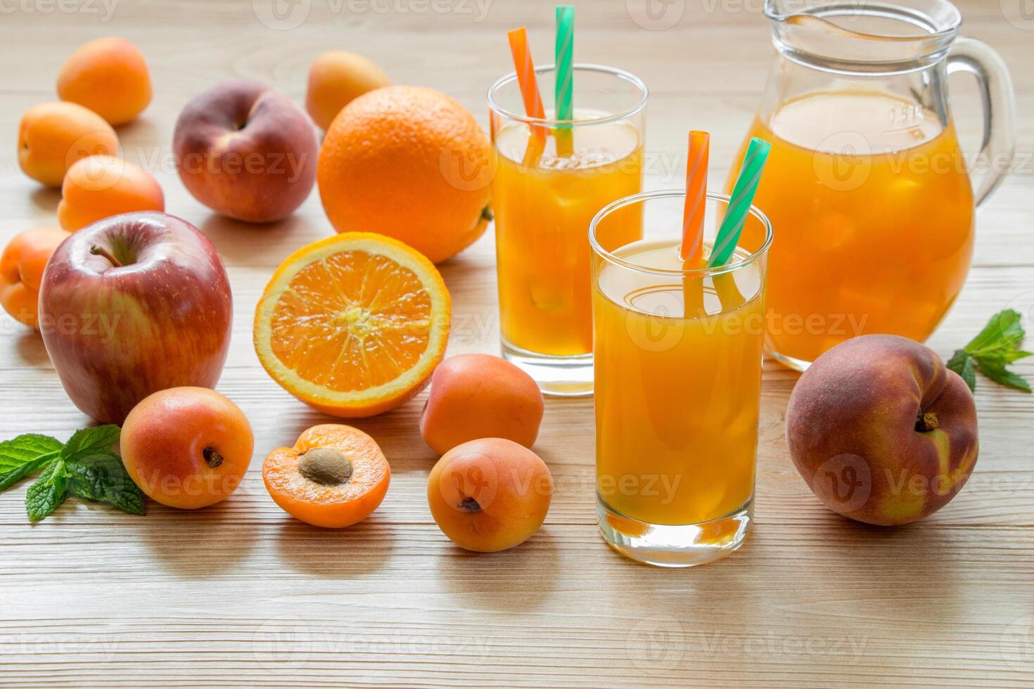 Fruit juice in glasses with ice near the jar of juice and scattered apricots, peaches, apple, orange on a light wooden background. Apricot peach apple orange juice with ice. Horizontal. photo