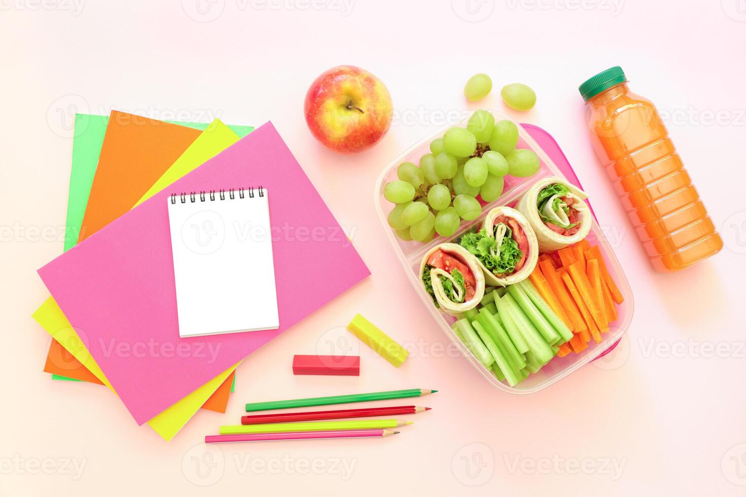 School supplies and lunch box with tasty rolls, cucumbers, carrots, grapes, apple, bottle of juice on light pink background, back to school concept. Horizontal. Top view. photo