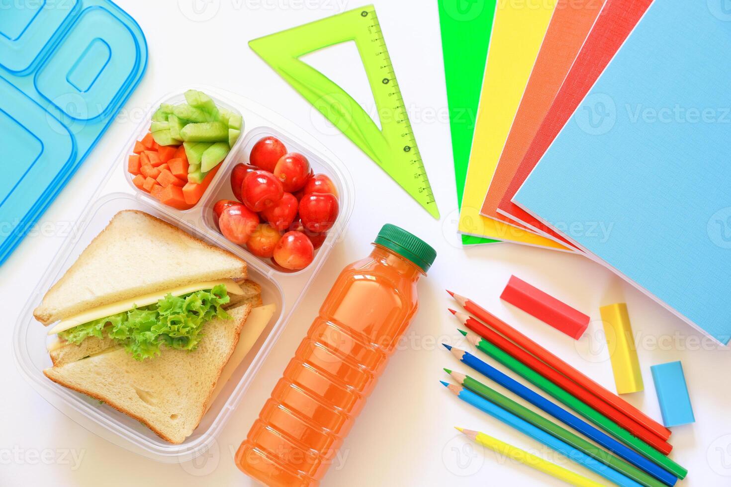 Variety of fresh snacks including sandwiches, fruits, and vegetables arranged neatly in lunch box alongside vibrant stationery, highlighting preparation for school day. photo