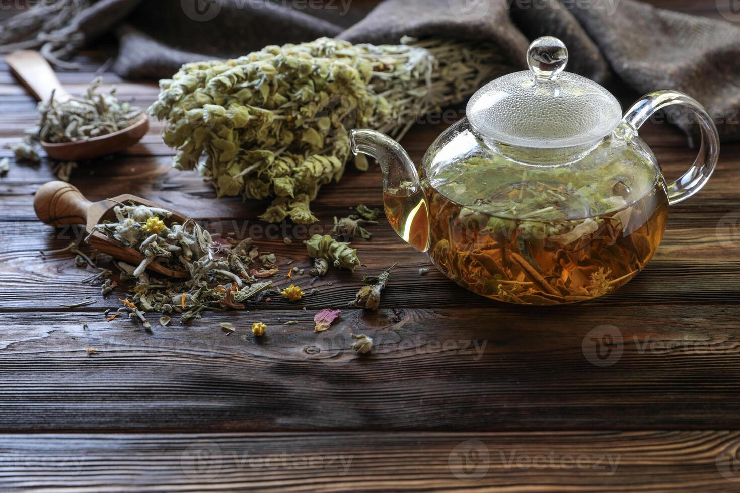 Tea in glass teapot, surrounded by fresh herbs and dried flowers on weathered wooden table, creating warm and inviting atmosphere for relaxation and enjoyment. photo