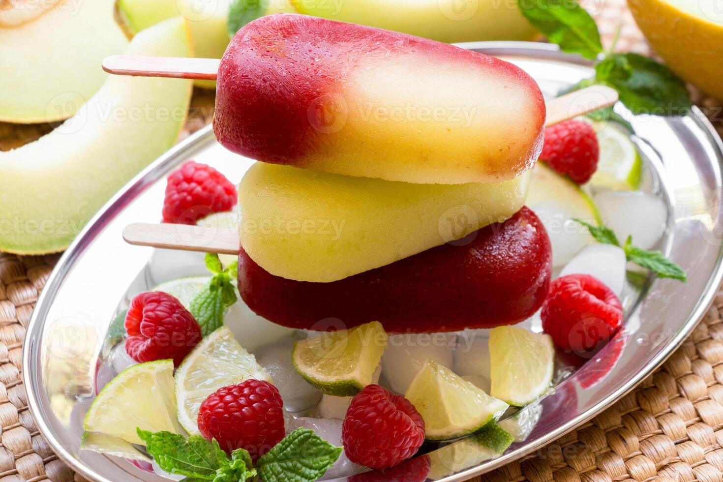 Vibrant melon raspberry ice cream sits atop a mound of ice cubes, surrounded by fresh raspberries, mint leaves, and sliced lime. photo