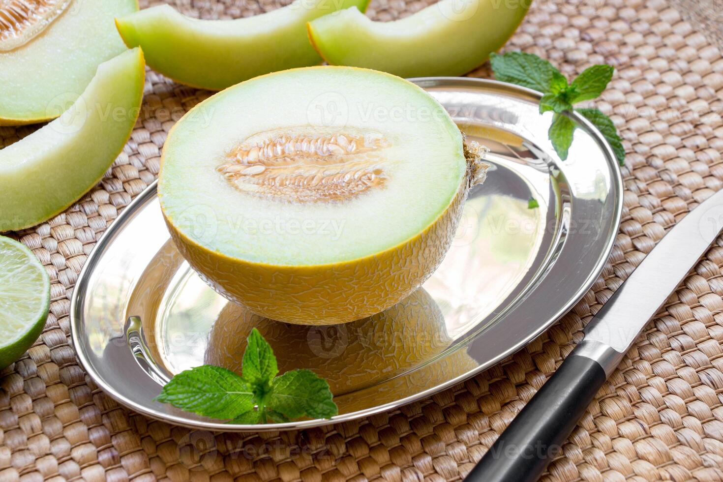 Diagonally at the center on a silver platter half of melon near melon slices, mint leaves, knife on a straw napkin background. Half of melon on silver platter. Horizontal. Close. photo