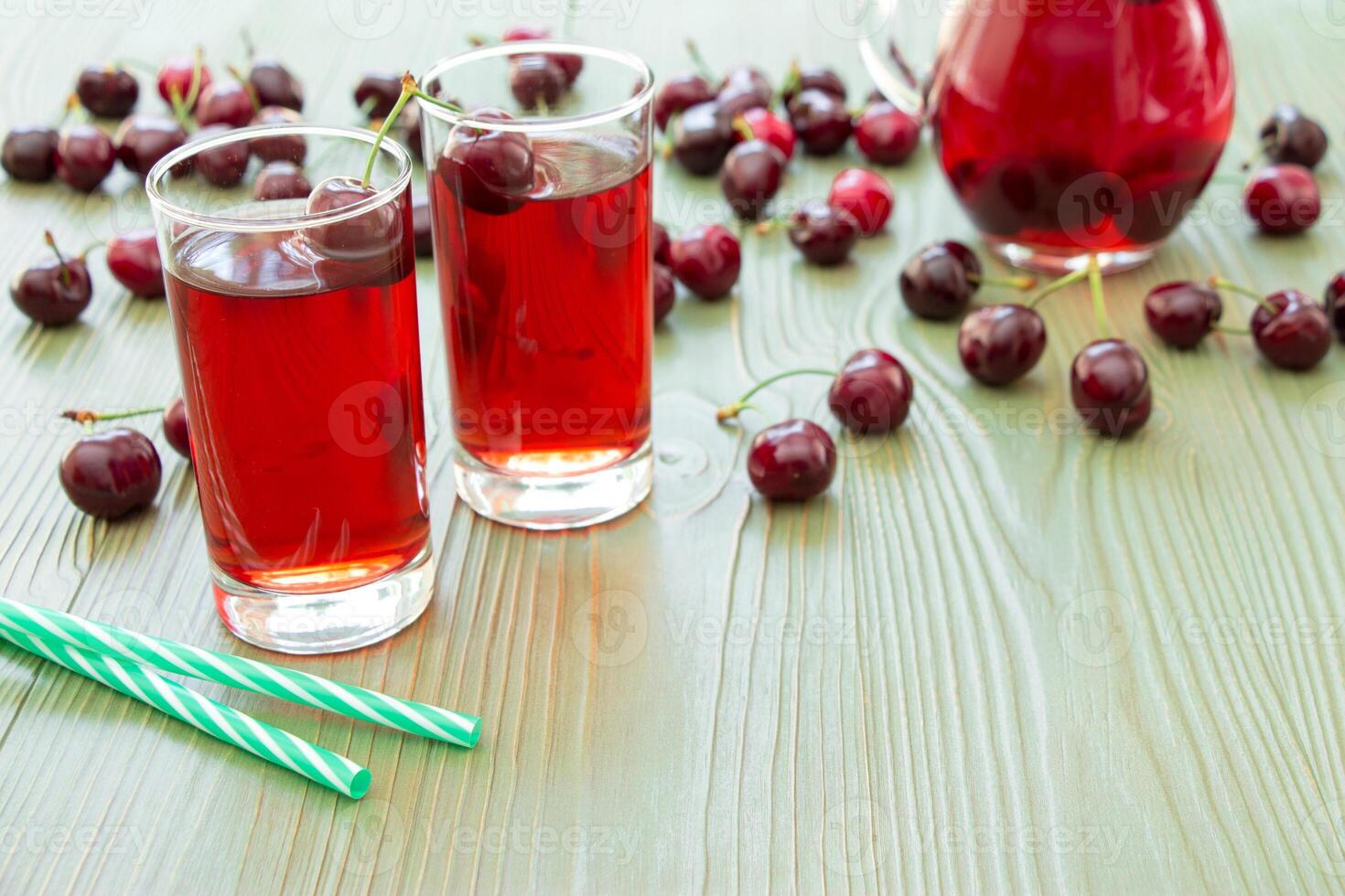 Two glasses of juice, green straws, scattered cherries, jar of cherry juice, on right empty space for text on green background. Cherries, cherry juice and empty space. Horizontal. photo
