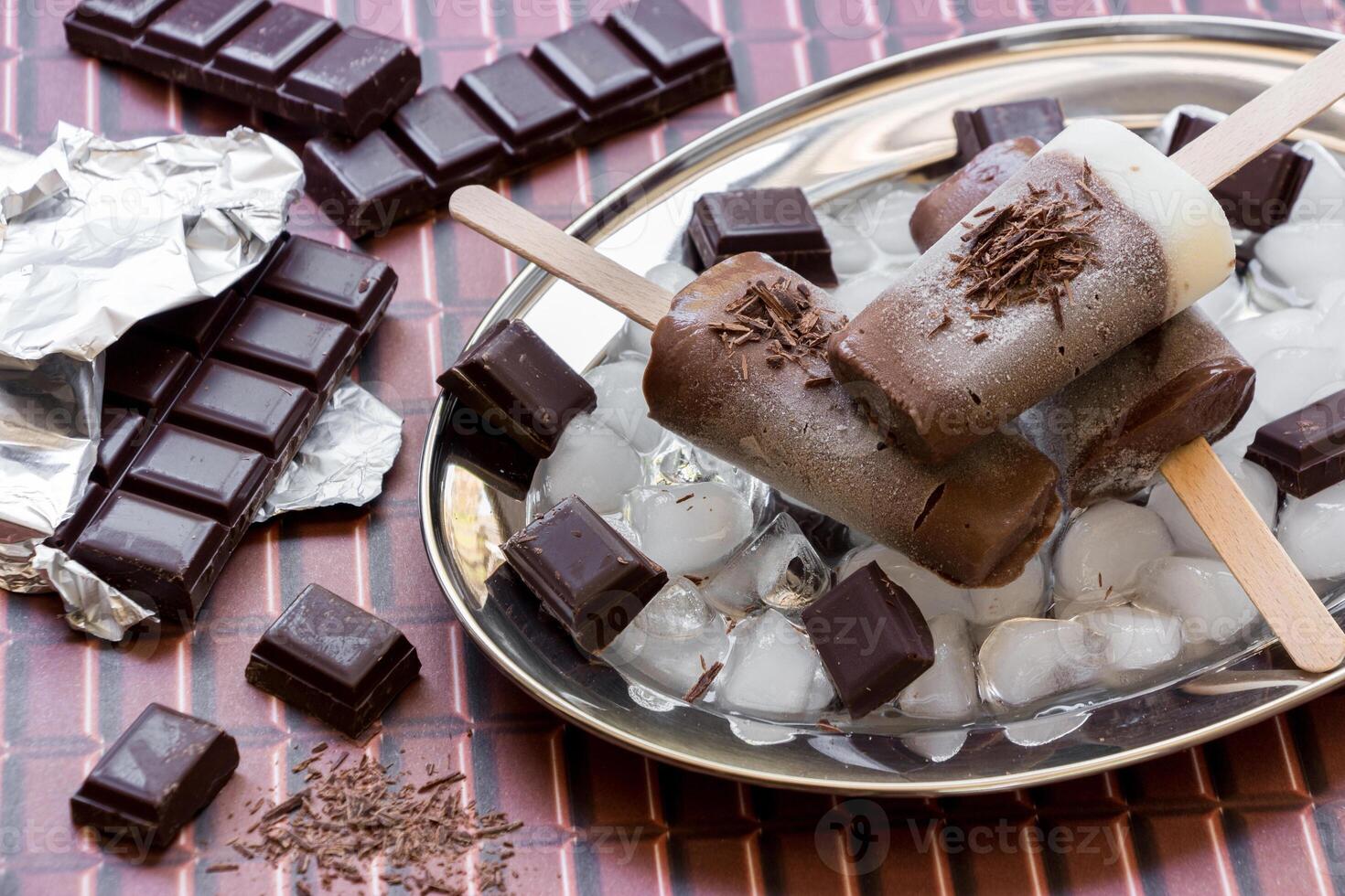 Chocolate vanilla ice cream on a stick on a silver platter with ice and pieces of chocolate near pieces of chocolate on a chocolate background. Chocolate vanilla ice cream popsicles. Horizontal. photo