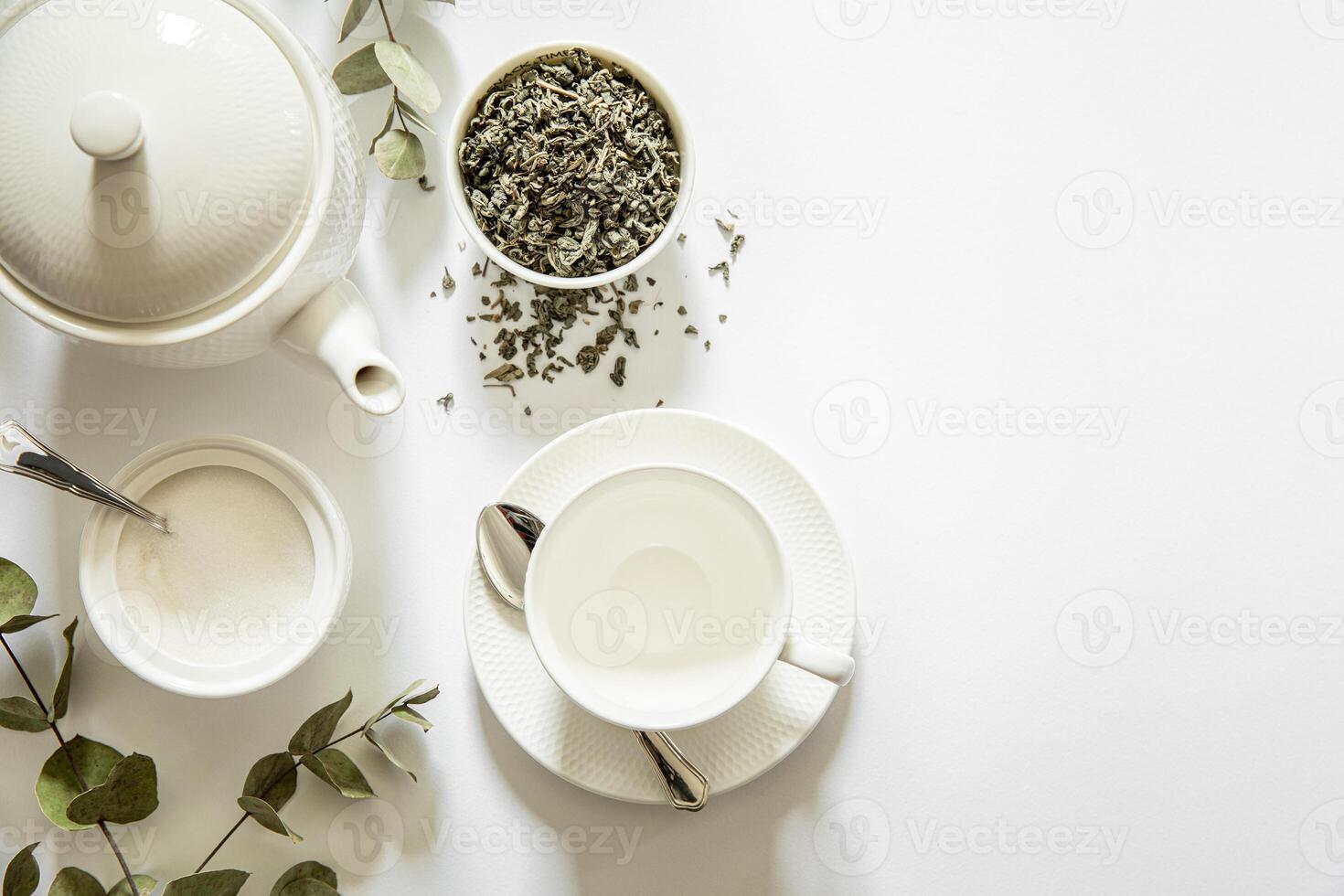 Teapot, cup, and plate arranged neatly with loose herbal tea, sugar bowl, and eucalyptus leaves on white surface, creating serene atmosphere for tea enjoyment. photo