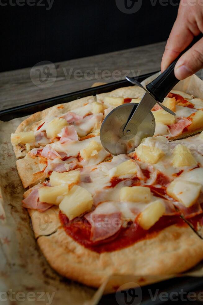 a person cutting a hawaiian pizza with a pizza cutter photo