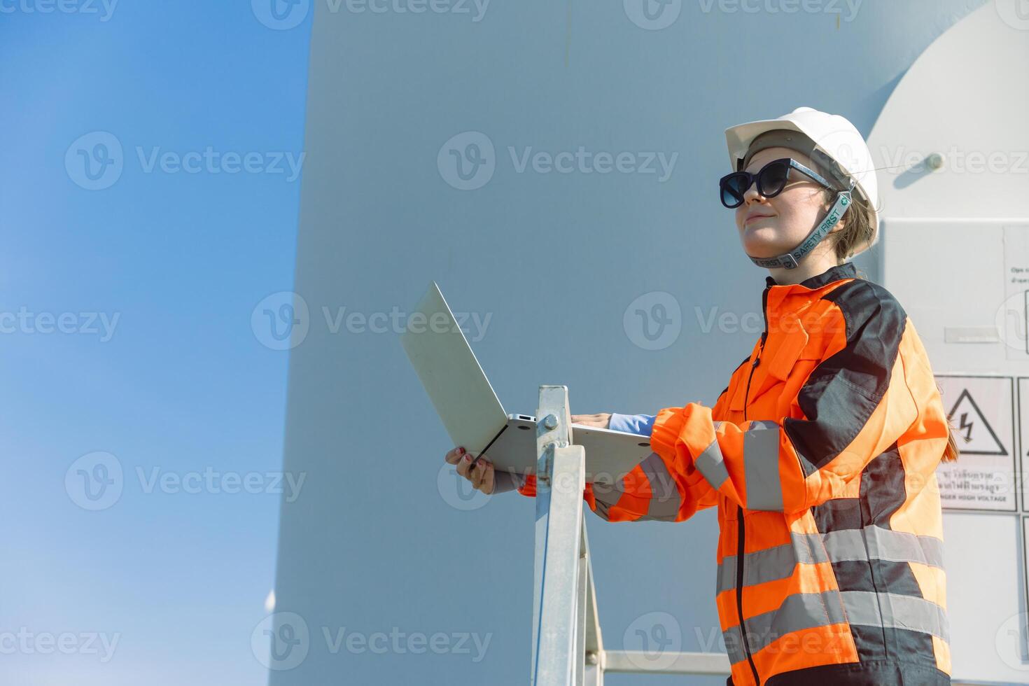 A woman in an orange and black jacket and safety glasses is standing on a ladder with a laptop photo
