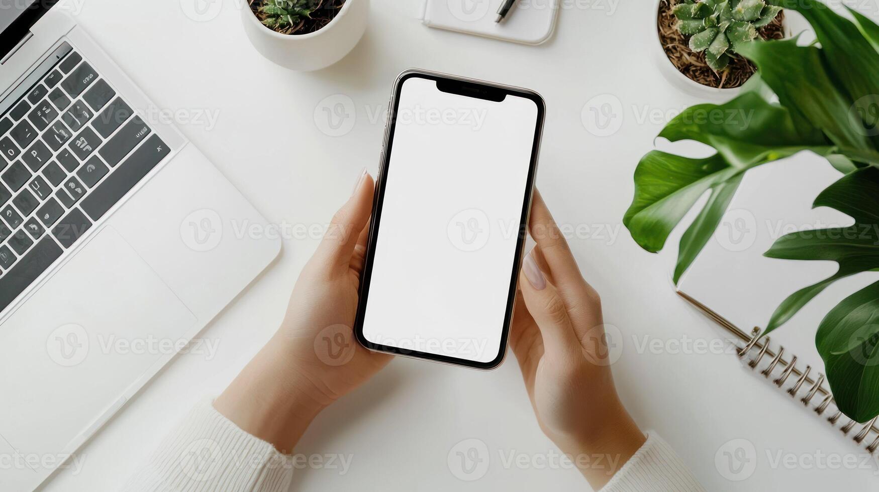 A person's hands holding a smartphone with a blank white screen, placed on a white desk with a laptop, a notebook, and a plant in the background. photo