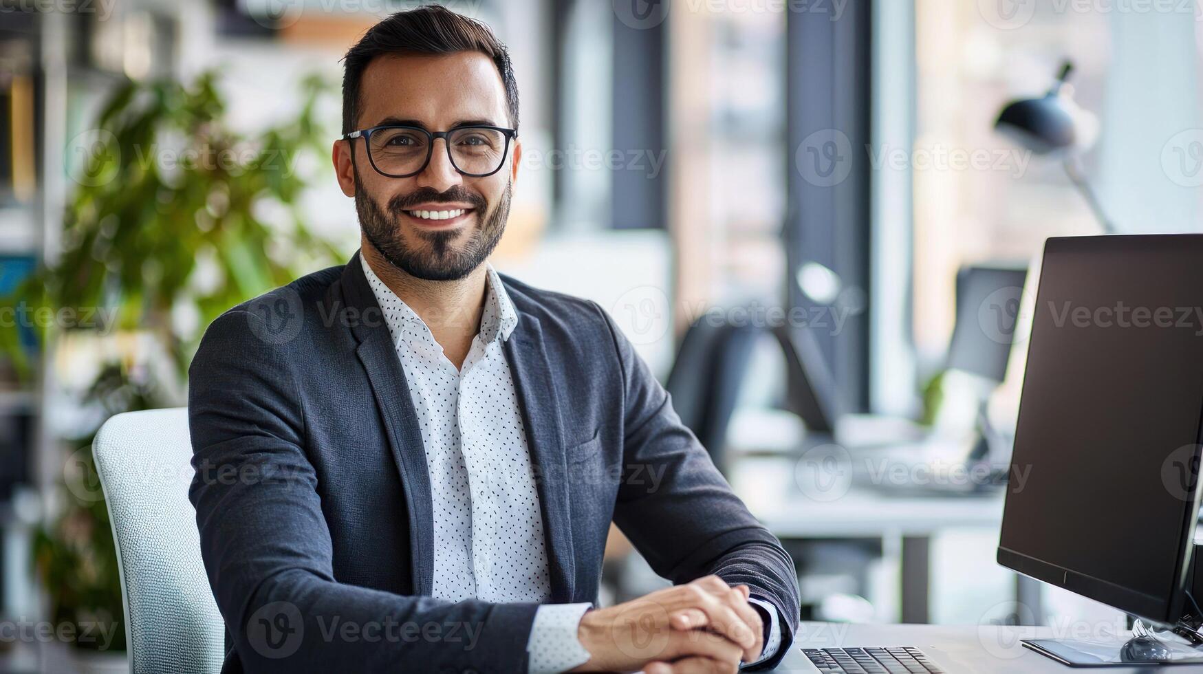 A smiling man in a suit sitting at a desk in an office with a computer monitor and a plant in the background. photo