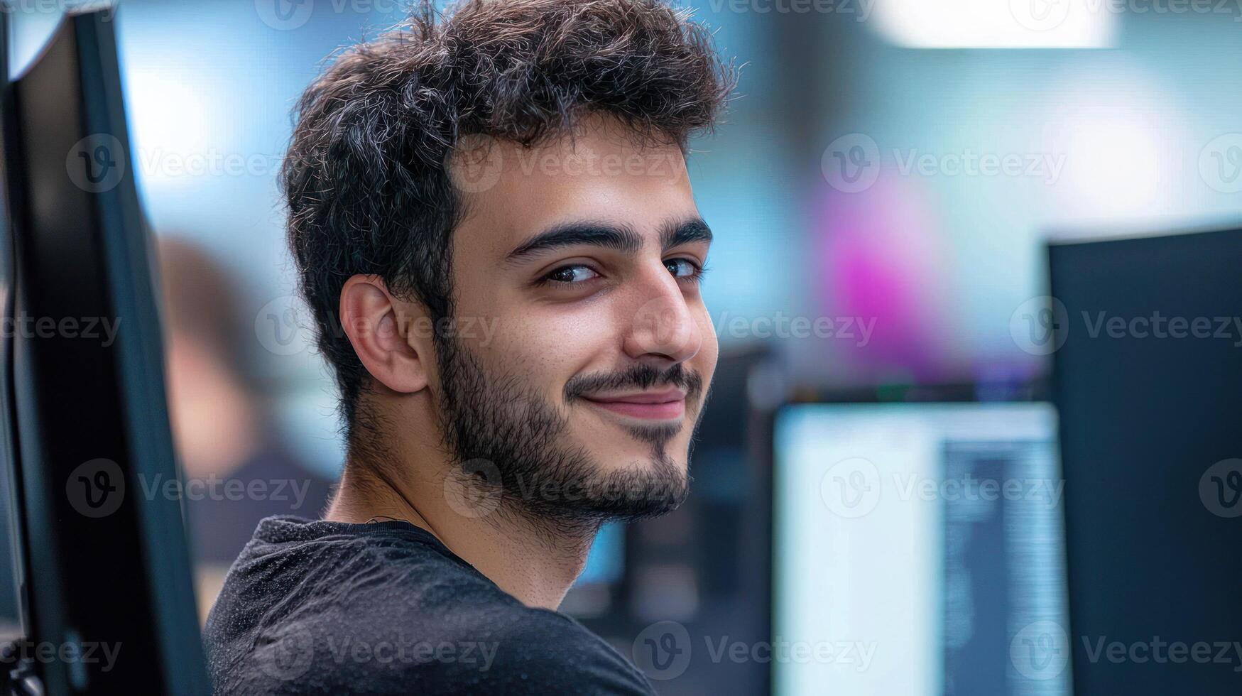 A young man with curly hair, wearing a black shirt, sitting in front of a computer in a modern office environment. photo