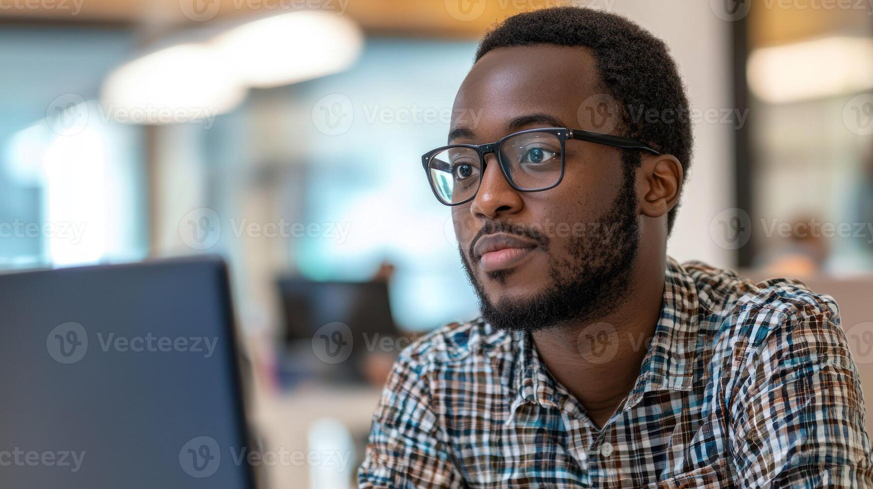 A young man wearing glasses, seated at a desk in an office environment, working on a computer. photo