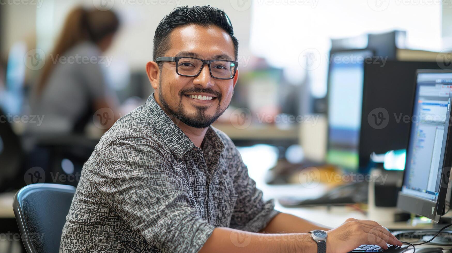 A smiling man in a casual office setting, wearing glasses, sitting at a desk with a computer, surrounded by office furniture and other employees in the background. photo