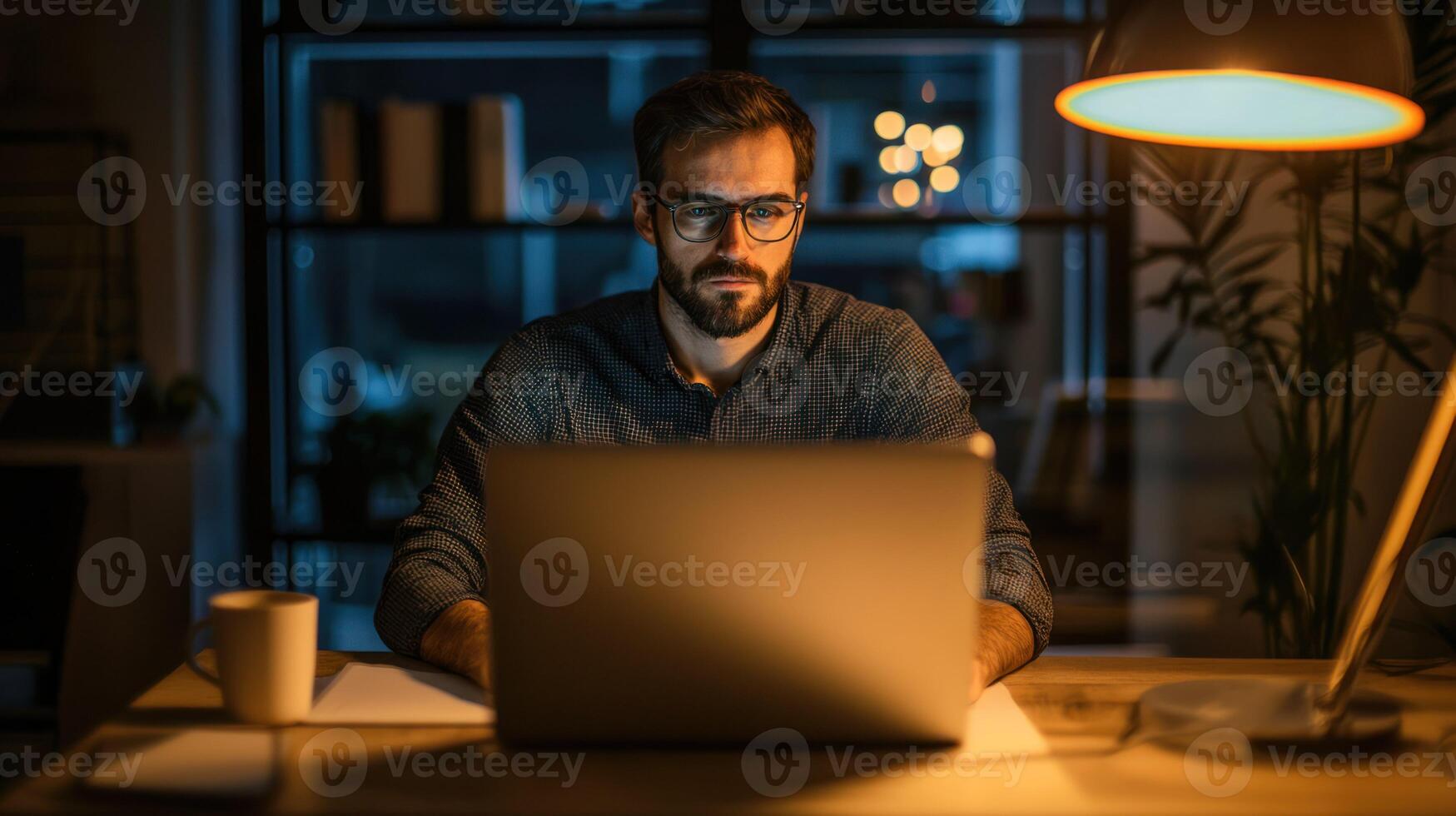 A man wearing glasses, sitting at a desk with a laptop, working late in a dimly lit office with a potted plant and a lamp. photo