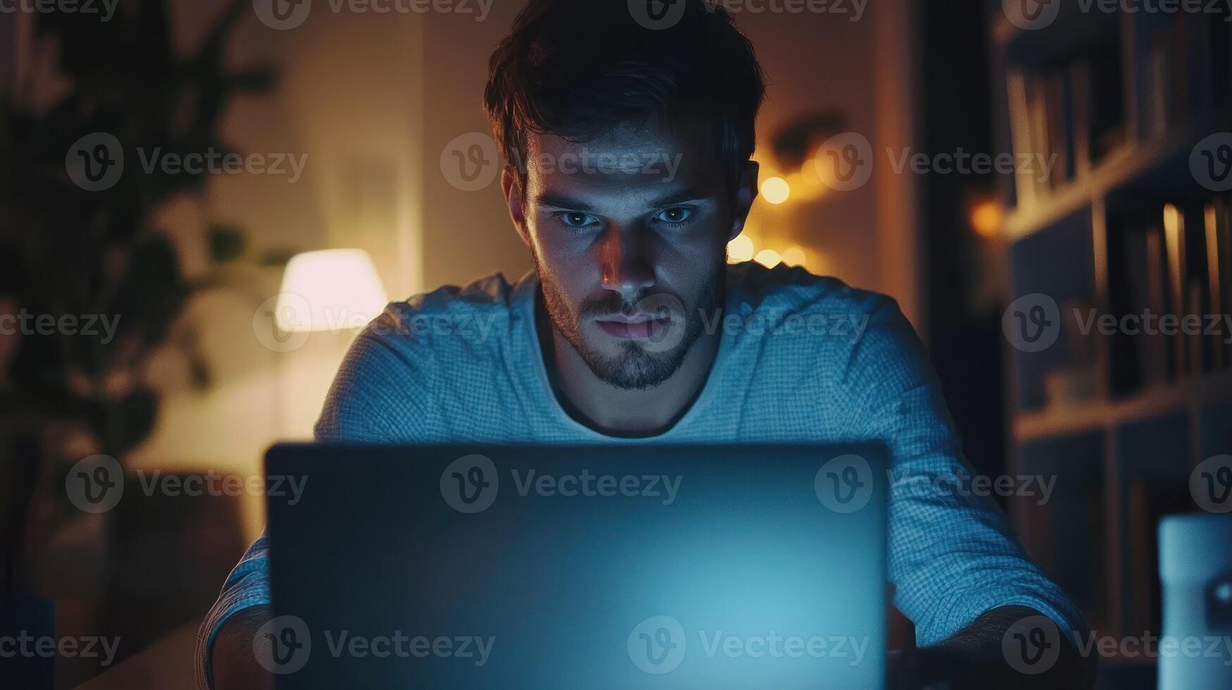 A man working late at night in a dimly lit room, focused on his laptop screen. photo