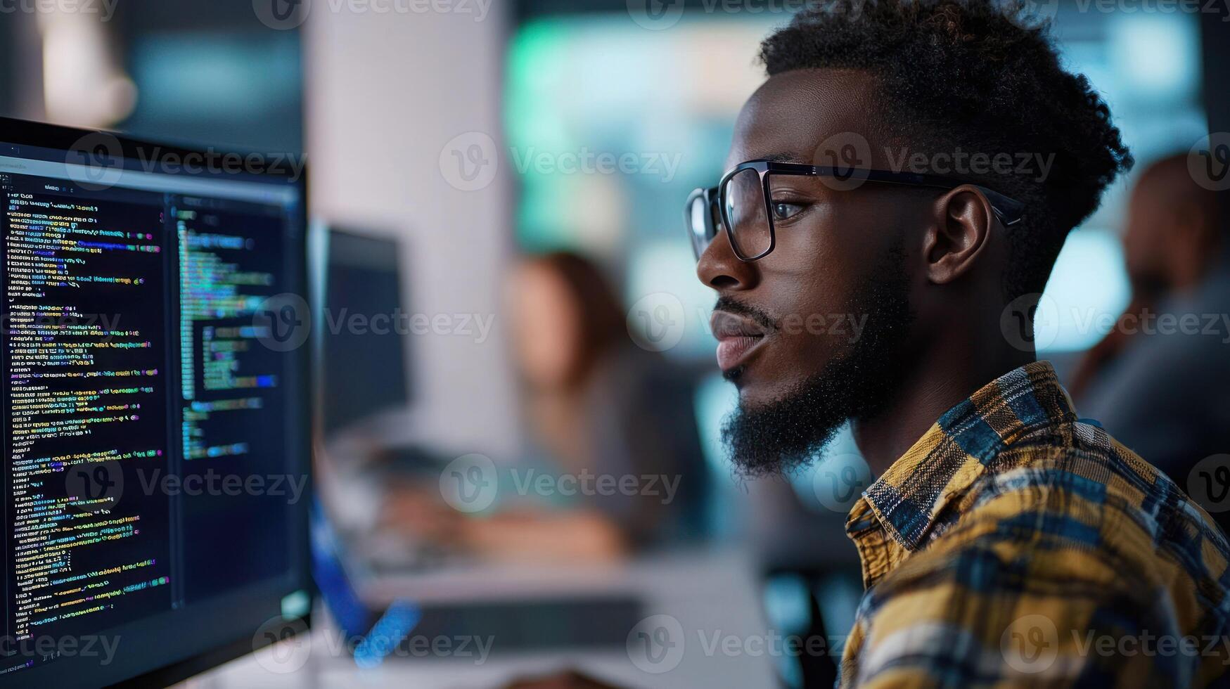 A young man wearing glasses, working on a computer in a busy office with other people in the background. photo