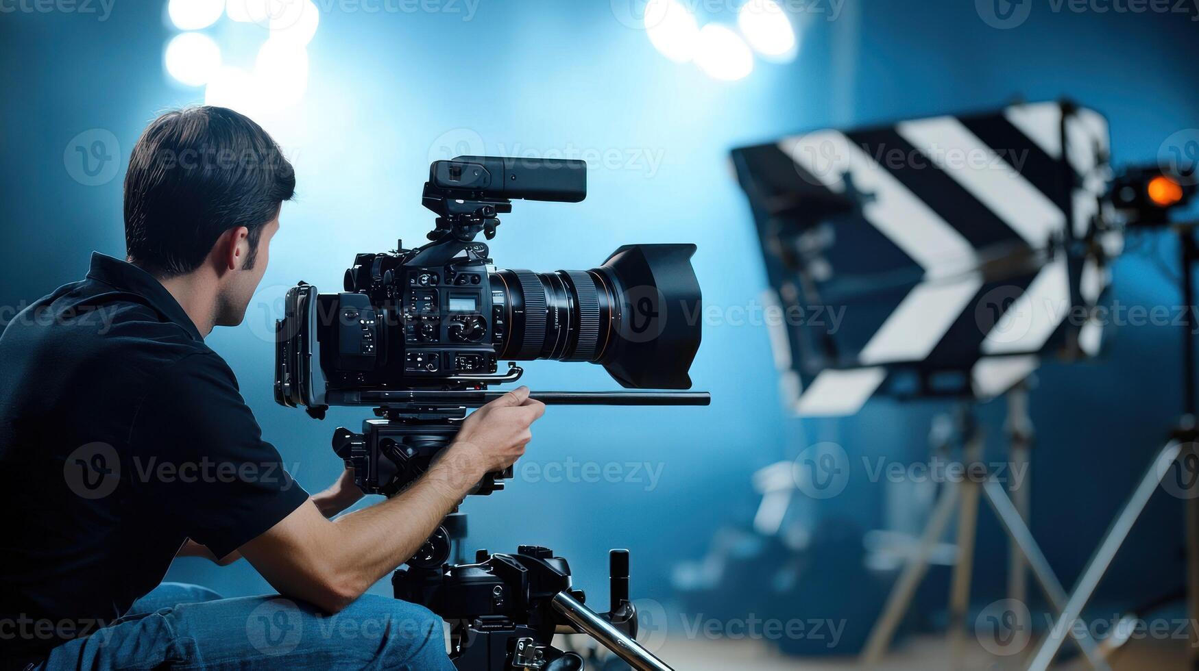 A man in a black shirt is kneeling on a blue mat, holding a large camera with a large lens, in front of a black and white striped backdrop with a green screen. photo