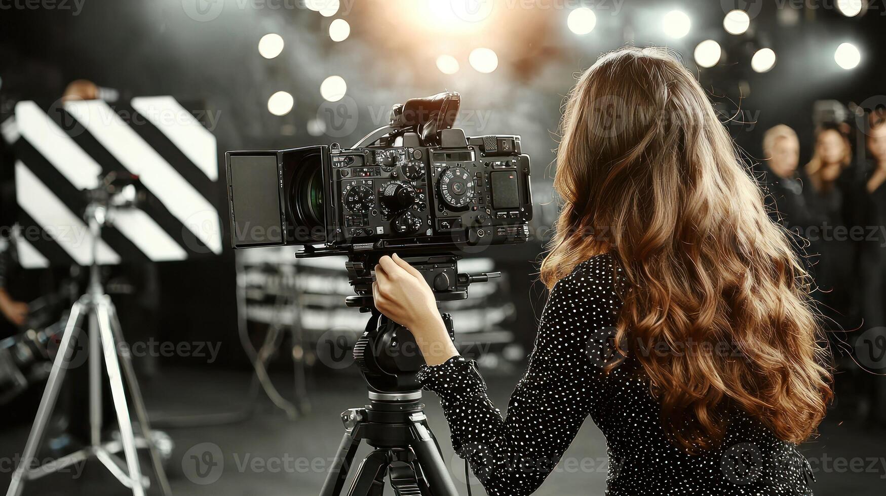 A woman with long, wavy hair standing in front of a camera on a stage with a microphone, surrounded by a blurred audience. photo