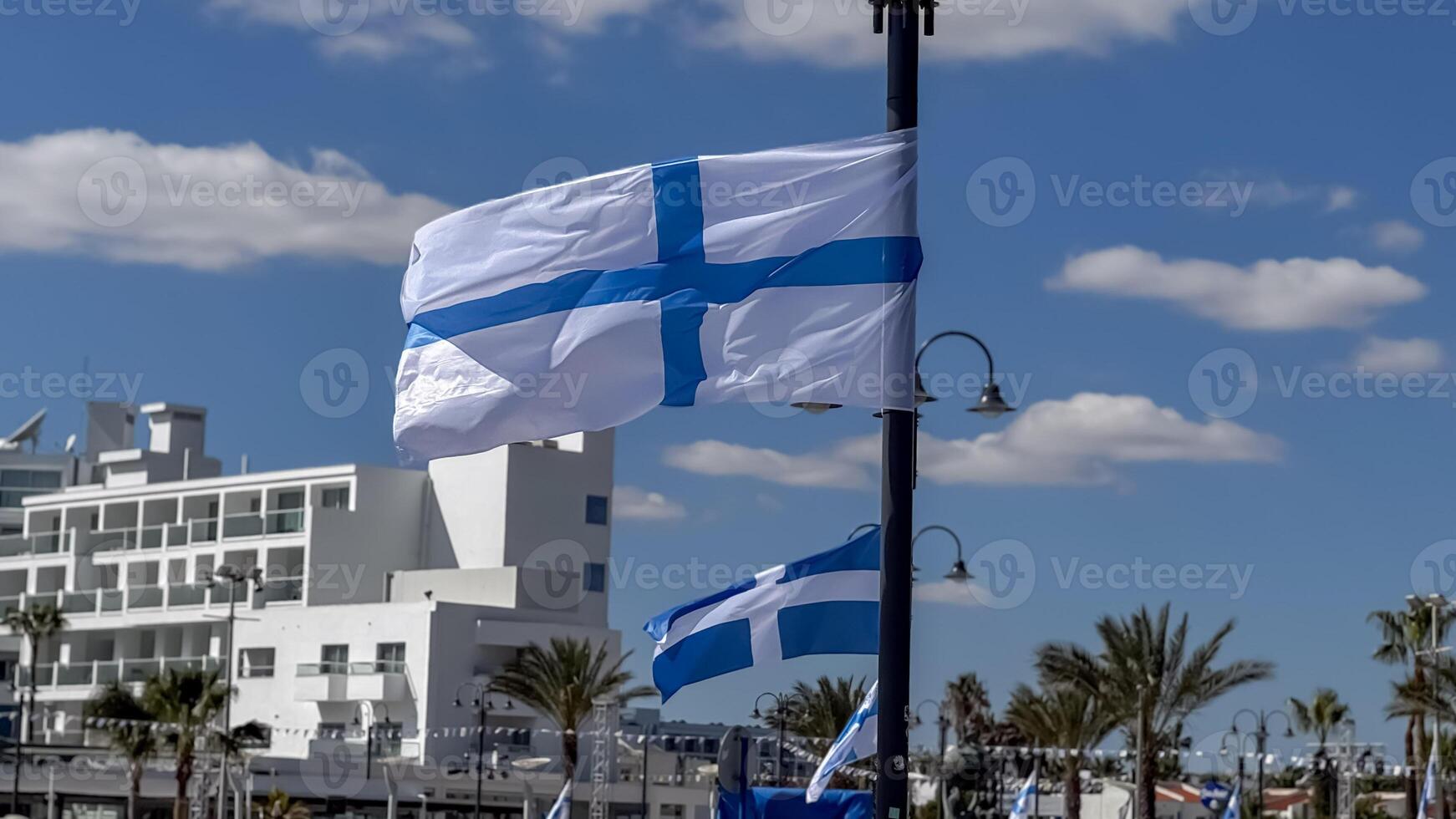 National flags of Greece and Cyprus blow in a strong breeze under a clear sky beside palm-lined coastal buildings. photo