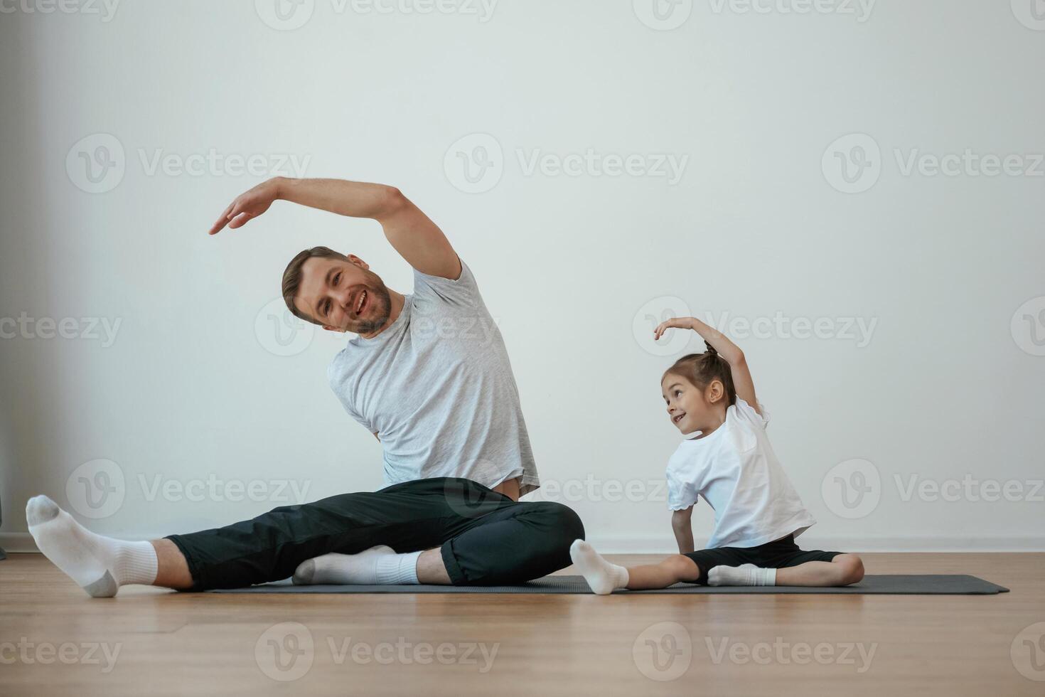 Against white ,wall, stretching. Father with little daughter are doing yoga at home photo