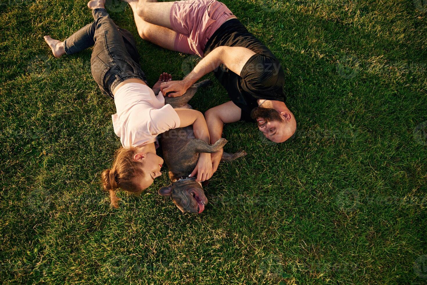 On, the grass, lying down. Man and woman are with dog on the field photo