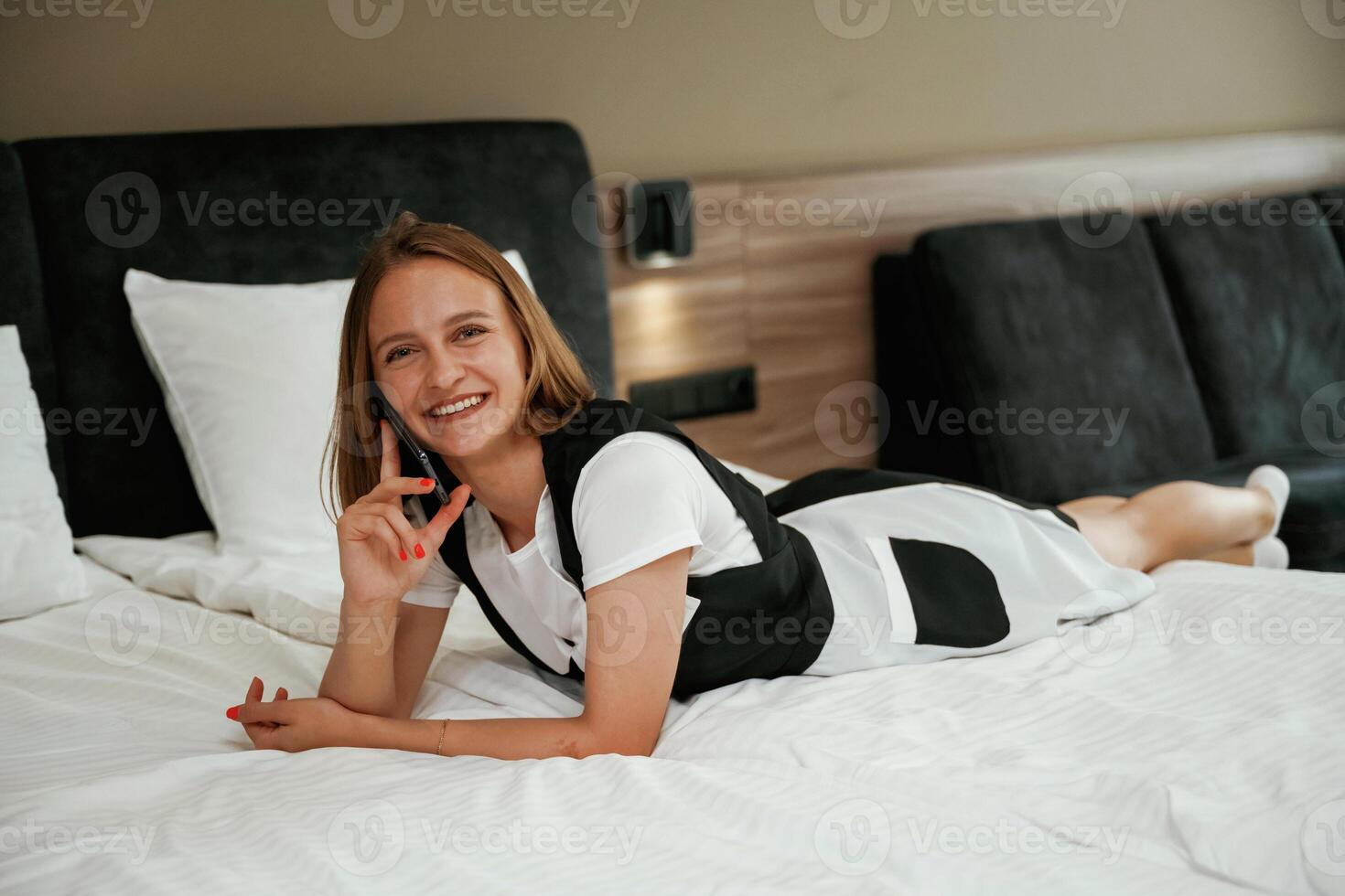 Cheerfu,l facia,l expression, lying down on bed with phone. Young chambermaid is in hotel room photo