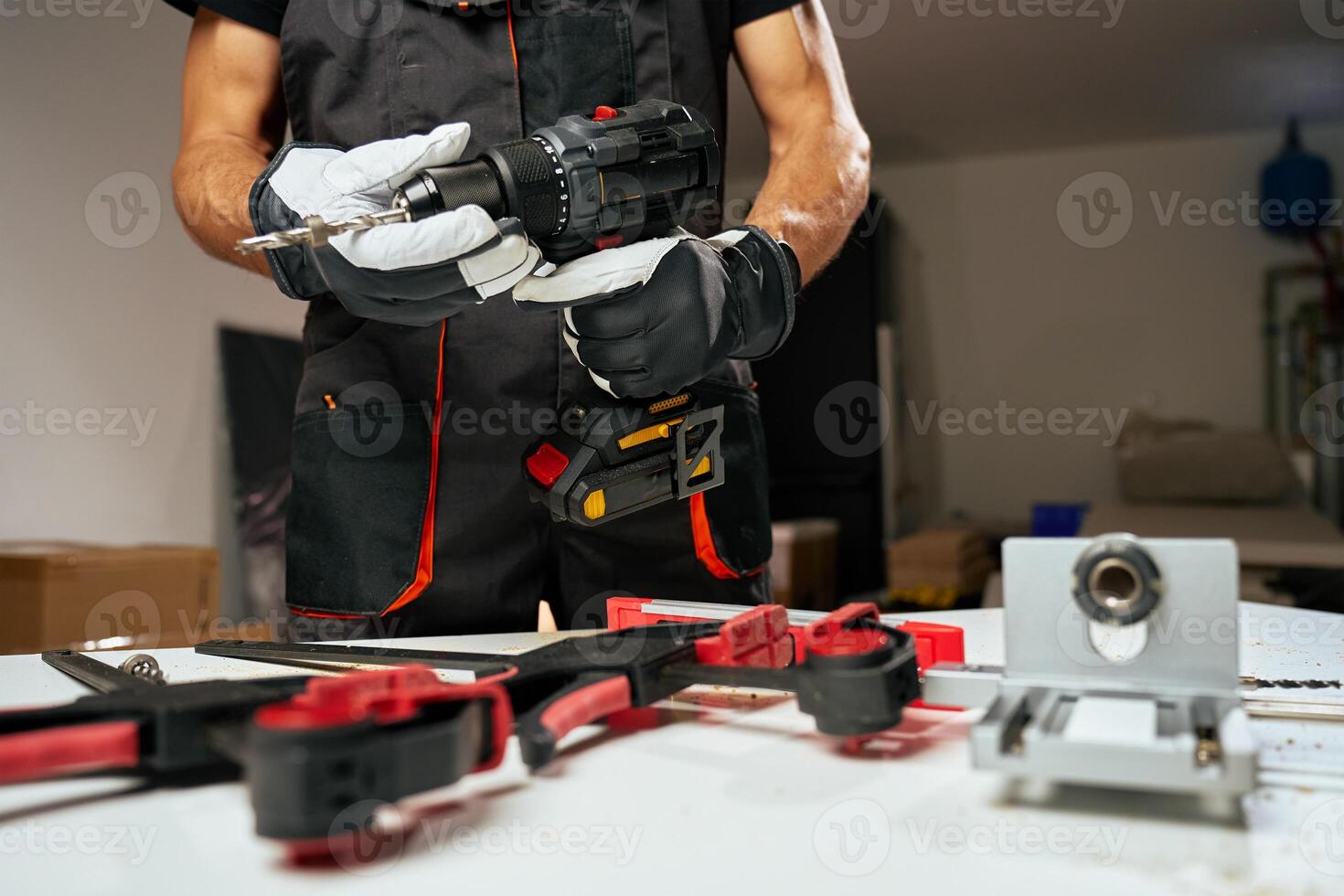 Person wearing safety gloves and work overalls holding cordless power drill over workbench with tools and components photo