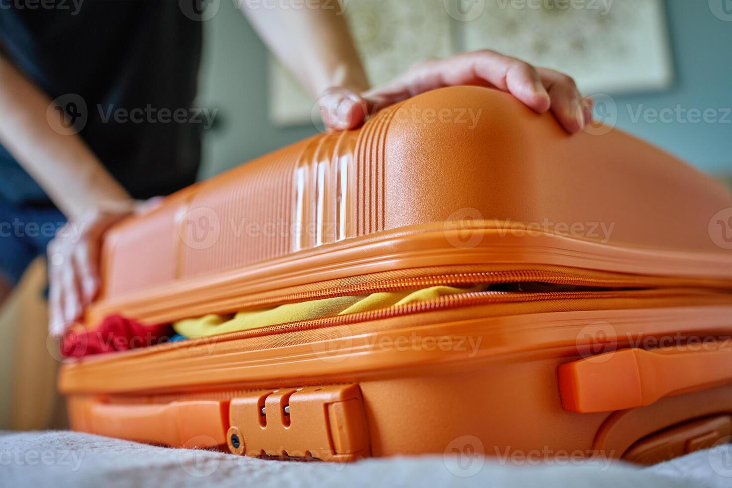 Woman trying to close suitcase at home. Female press down overfilled orange suitcase while packing clothes. Person preparing for departure photo