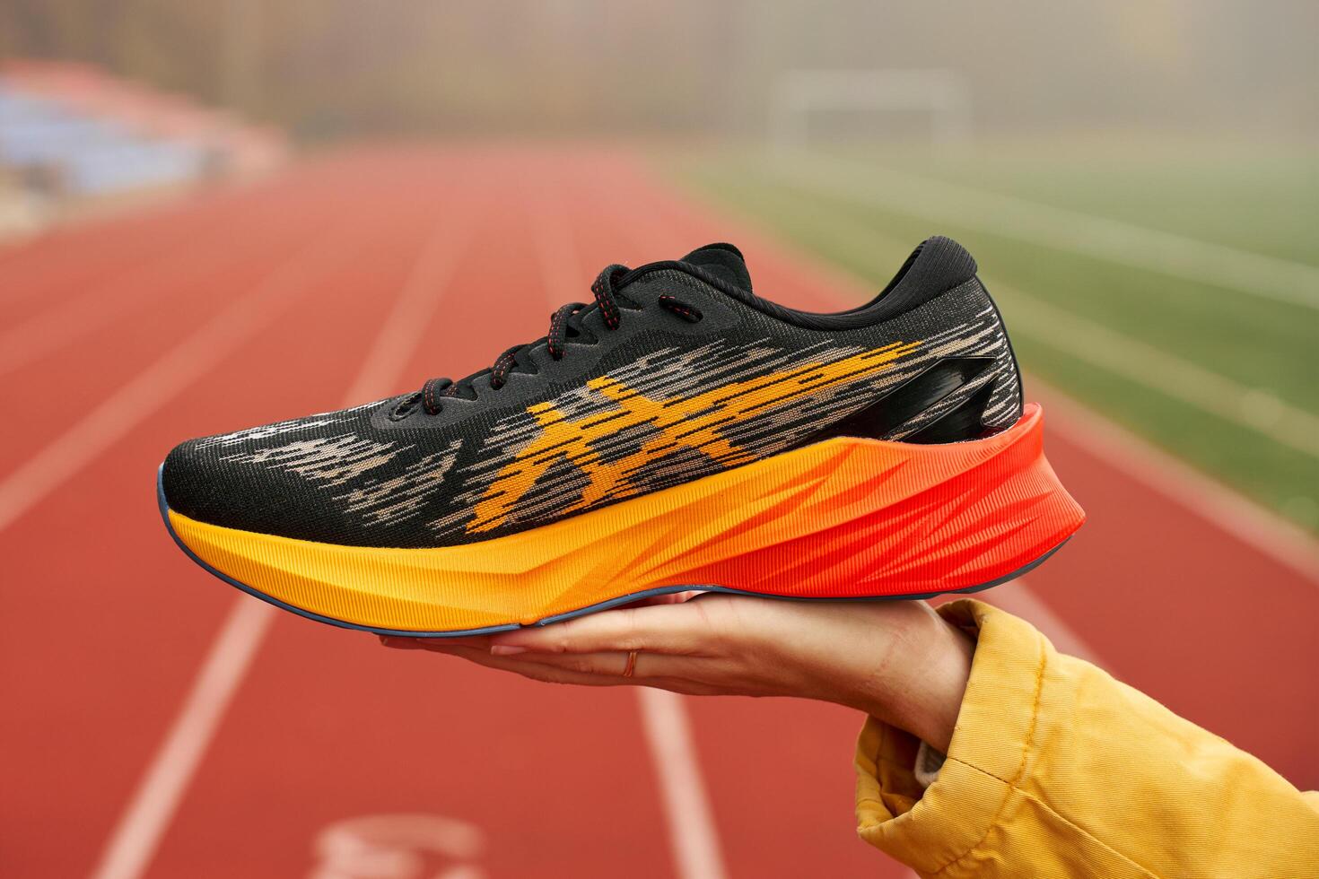 Black and orange Asics Novablsat 3 running shoe is held in hand against blurred background of track and field. Sport exercises and an active lifestyle concept. Wroclaw, Poland - November 6, 2024 photo