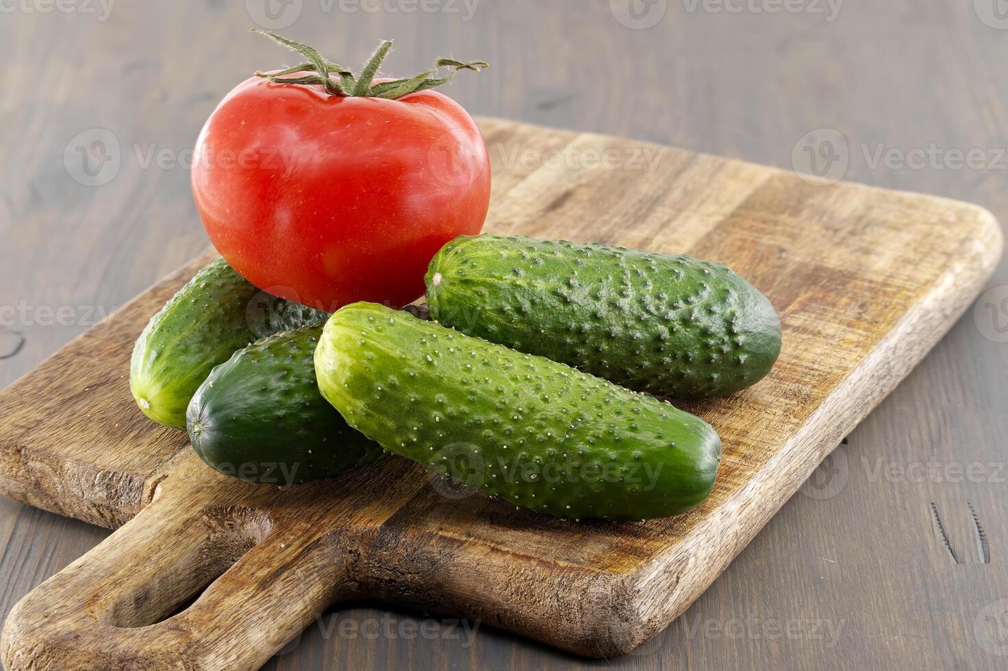 Fresh cucumbers and tomato lying on wooden cutting board photo