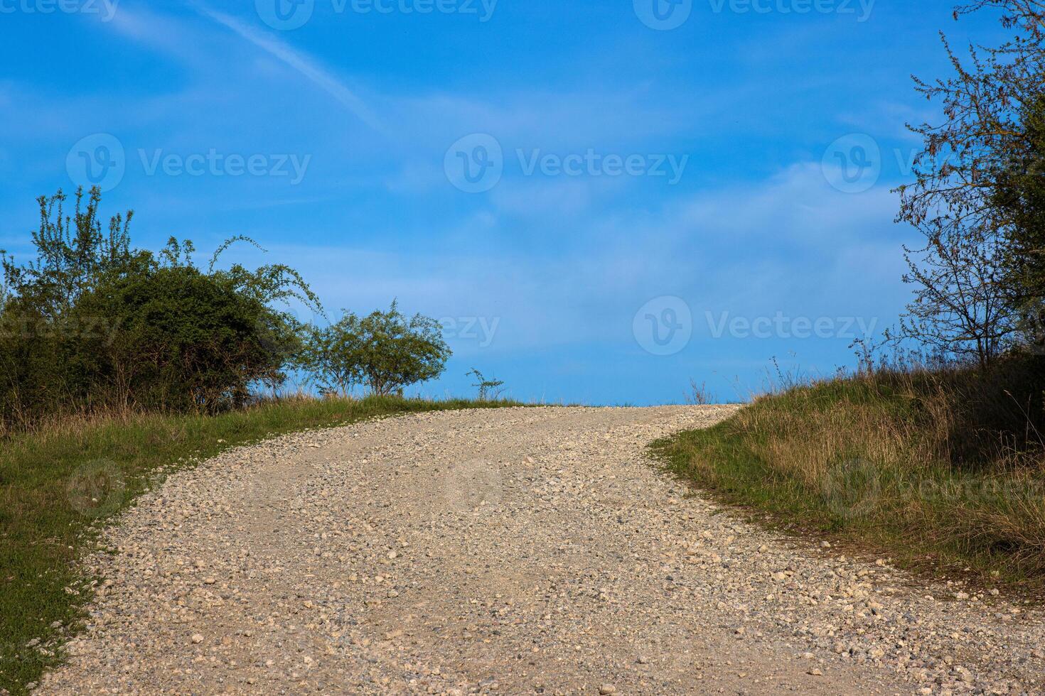 An empty gravel road winds uphill through a rural landscape. The path leads toward the horizon under a blue sky with soft clouds 18 photo